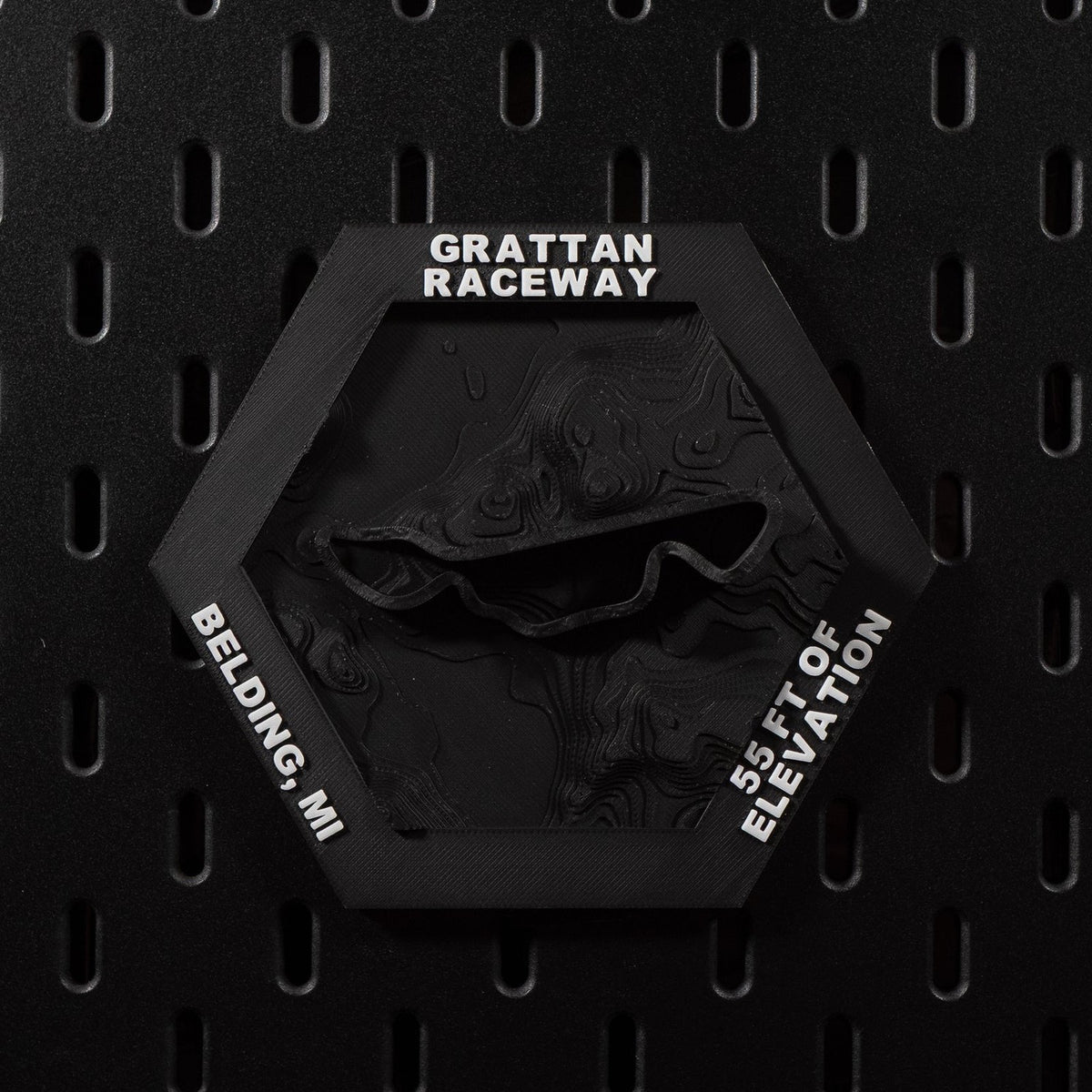 Grattan Raceway hex mounted flat on a black pegboard wall, all black with the circuit loop raised in relief